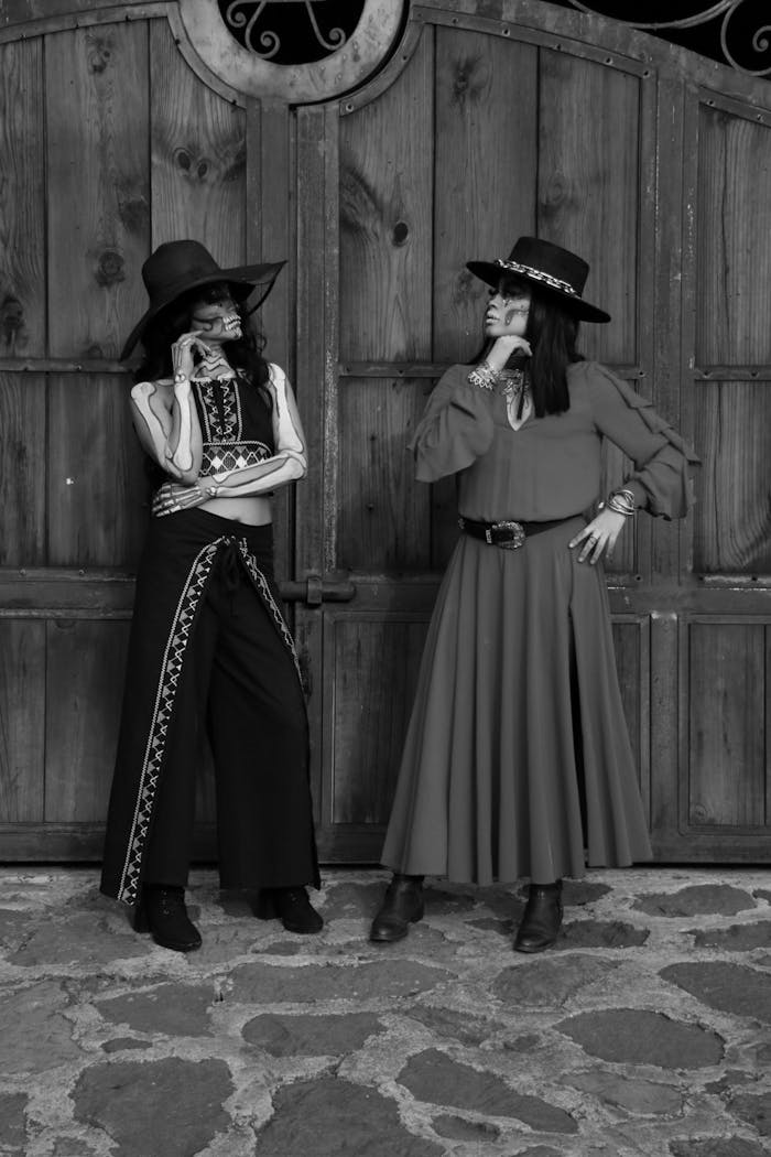 Two women in vintage western outfits pose against a rustic wooden gate.