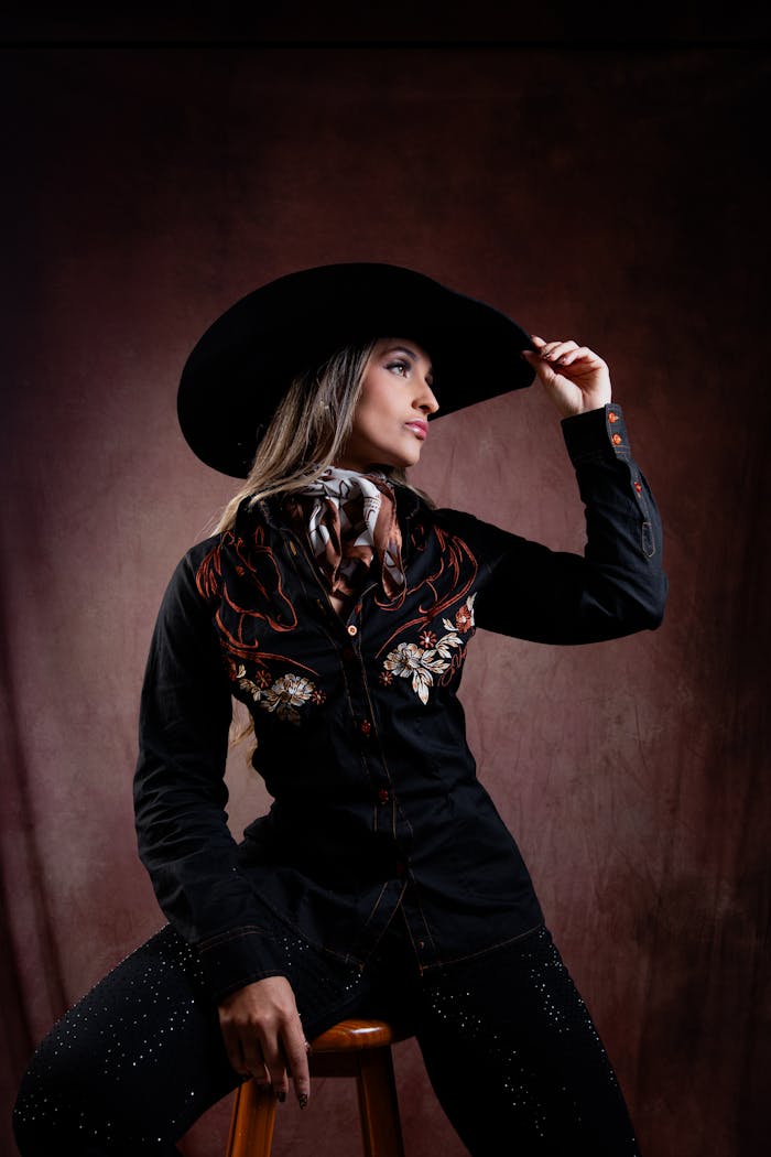 Home Stylish woman in western attire and cowboy hat on a stool, exuding confidence.